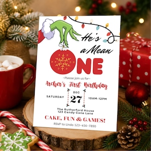 May include: A white rectangular birthday invitation card featuring a green hand holding a red ornament with a gold snowflake. The text reads He's a Mean ONE, Archer's First Birthday, Dec 27, 10AM-12PM, The Rutherford House, 123 Candy Cane Lane, CAKE, FUN & GAMES!, and RSVP to Linda 123-456-7890.