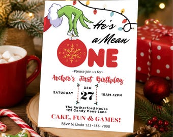 May include: A white rectangular birthday invitation card featuring a green hand holding a red ornament with a gold snowflake. The text reads He's a Mean ONE, Archer's First Birthday, Dec 27, 10AM-12PM, The Rutherford House, 123 Candy Cane Lane, CAKE, FUN & GAMES!, and RSVP to Linda 123-456-7890.