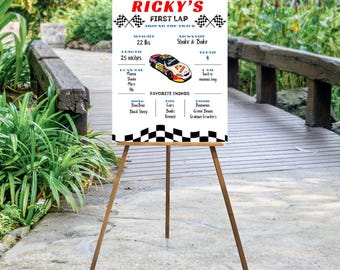 May include: A wooden easel displaying a personalized sign for "Ricky's First Lap." The sign features a race car graphic, checkered flags, and details like weight (22 lbs), length (25 inches), and favorite things. The background shows a wooden bridge and greenery.