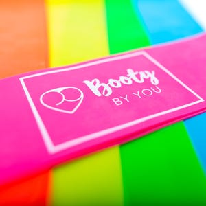 Könnte beinhalten: Ein Set aus fünf Widerstandsbändern in verschiedenen Farben, darunter Pink, Orange, Gelb, Grün und Blau. Das rosa Band hat den Text "Booty By You" aufgedruckt.