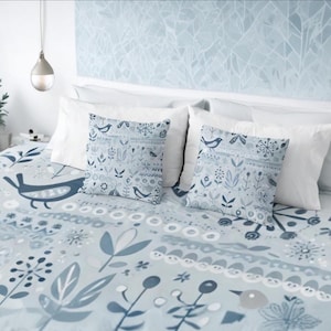 Pode incluir: Uma cama com uma colcha estampada floral azul claro e duas almofadas a condizer. As almofadas e a colcha apresentam um design de pássaros e flores em tons de azul e branco. Almofadas brancas e uma cabeceira branca completam o conjunto de roupa de cama.