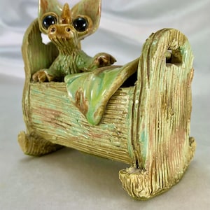 May include: A ceramic sculpture of a creature with large black eyes and a horn, in a green and brown rocking cradle. The creature has green fur and is lying on a green blanket. The cradle has a textured wood-like appearance, with a rustic finish.
