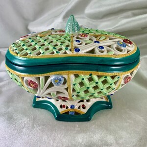 Puede incluir: Un tazón de servir de cerámica blanca con tapa, que presenta un diseño de celosía verde, amarillo y blanco con acentos florales. El tazón tiene una base festoneada y una perilla decorativa en la tapa.