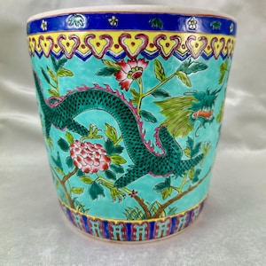 Puede incluir: Una maceta de cerámica turquesa con un diseño detallado de dragón y flores. La maceta presenta un dragón verde, flores rosas y un borde decorativo con detalles amarillos y azules. El diseño general es ornamentado y colorido.