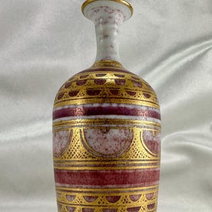 May include: A white ceramic vase with a narrow neck and a wide base. The vase is decorated with gold and red geometric patterns.