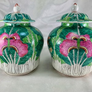 May include: Two matching ceramic jars with lids. The jars are painted with a vibrant design of pink flowers and green leaves against a green background. The lids are also green with white finials. The jars have a bulbous shape and are set on a white surface.