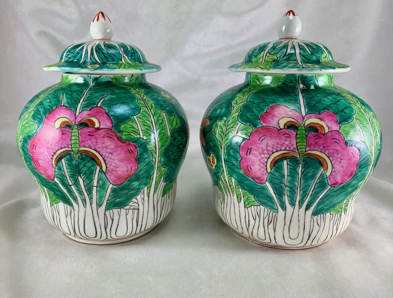 May include: Two matching ceramic jars with lids. The jars are painted with a vibrant design of pink flowers and green leaves against a green background. The lids are also green with white finials. The jars have a bulbous shape and are set on a white surface.