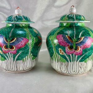 May include: Two matching ceramic jars with lids. The jars are green with a butterfly and leaf design. The butterfly has pink, blue, and yellow wings. The lids are green with a white finial. The jars are approximately 8 inches tall.