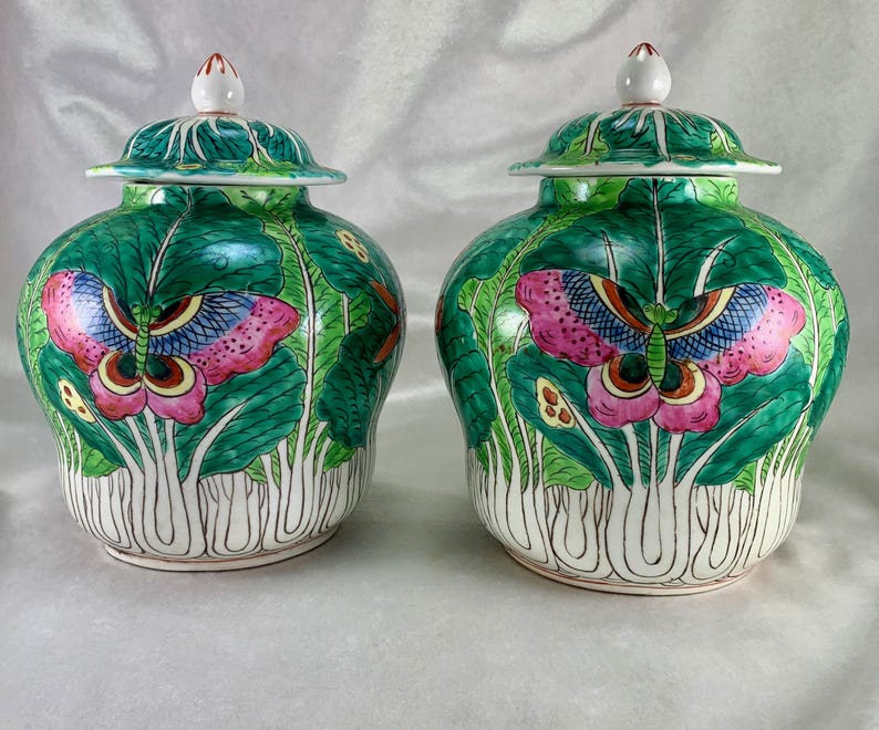 May include: Two matching ceramic jars with lids. The jars are green with a butterfly and leaf design. The butterfly has pink, blue, and yellow wings. The lids are green with a white finial. The jars are approximately 8 inches tall.