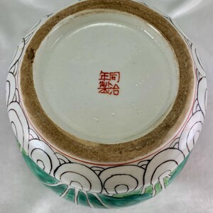May include: A ceramic vase with a wide opening, featuring a design of white, black, and green swirling patterns. The base has a red stamp with Chinese characters. The rim is brown with a red accent.