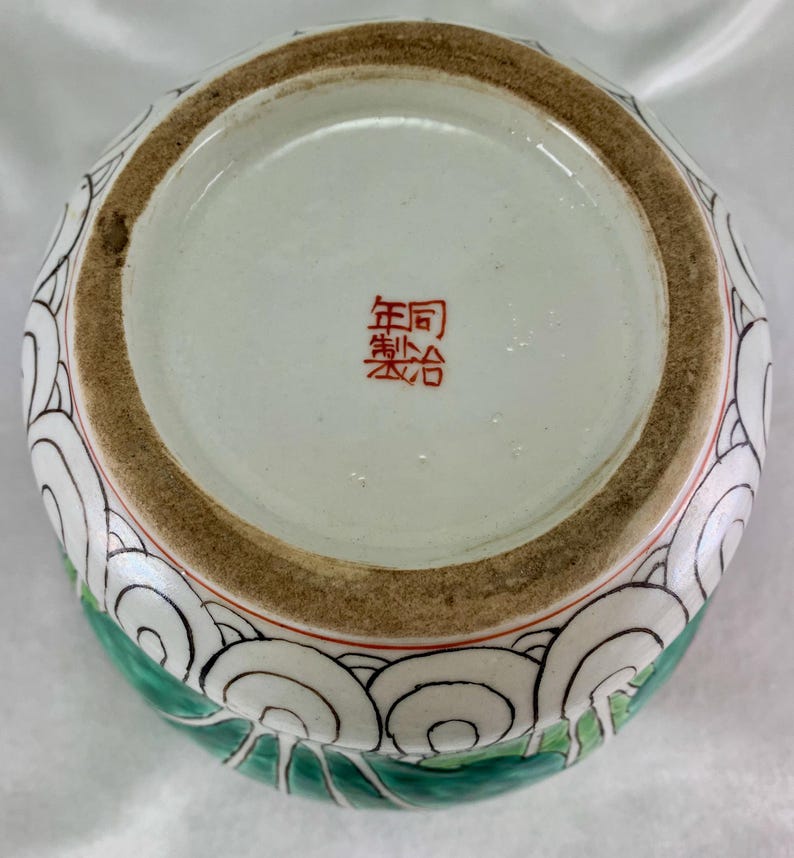 May include: A ceramic vase with a wide opening, featuring a design of white, black, and green swirling patterns. The base has a red stamp with Chinese characters. The rim is brown with a red accent.