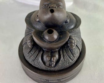 May include: A brown ceramic incense burner shaped like a smiling Buddha holding a round incense dish. The burner has a textured surface and a small hole on the top for incense sticks.