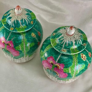 May include: Two matching, hand-painted ceramic jars with lids. The jars are a vibrant green with pink floral designs and white accents. The lids feature a white and green pattern with a small, red-tipped finial. The jars are likely decorative or for storage.