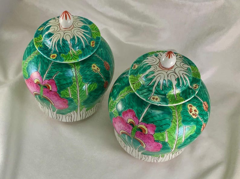 May include: Two matching, hand-painted ceramic jars with lids. The jars are a vibrant green with pink floral designs and white accents. The lids feature a white and green pattern with a small, red-tipped finial. The jars are likely decorative or for storage.