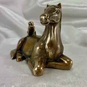 May include: A bronze sculpture of a horse in a seated position with a small monkey riding on its back. The sculpture has a detailed design and a polished finish. The horse is a light brown colour.