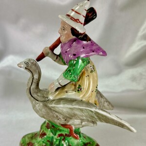 May include: A vintage ceramic figurine depicting a woman riding a goose. The woman wears a white hat with red stripes, a purple cape, and a yellow dress. The goose is gray, and the base is green with red accents. The figurine is hand-painted.