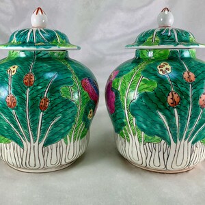 May include: Two matching green ceramic jars with lids. The jars are decorated with a leaf and insect design. The lids are white with a red tip. The jars have a bulbous shape and are set against a white background.