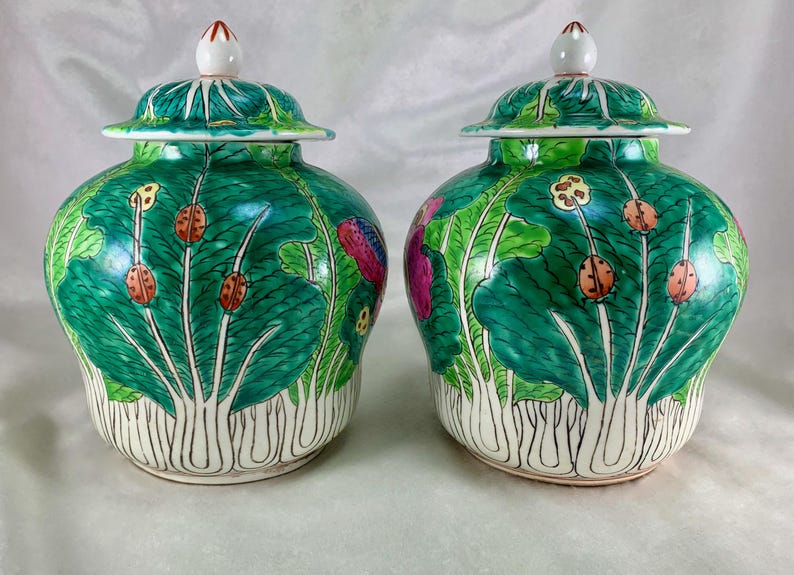 May include: Two matching green ceramic jars with lids. The jars are decorated with a leaf and insect design. The lids are white with a red tip. The jars have a bulbous shape and are set against a white background.