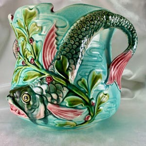 Puede incluir: Una jarra de cerámica turquesa con un diseño de pez. El pez es verde, rosa y blanco con un patrón de escamas detallado. La jarra tiene un asa con forma de cola de pez.