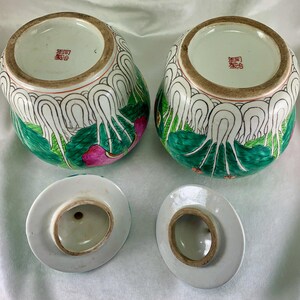 May include: Two antique, hand-painted ceramic jars with lids. The jars are decorated with green, white, and pink floral designs. The lids are white with a brown rim. The jars have a Chinese inscription on the bottom.