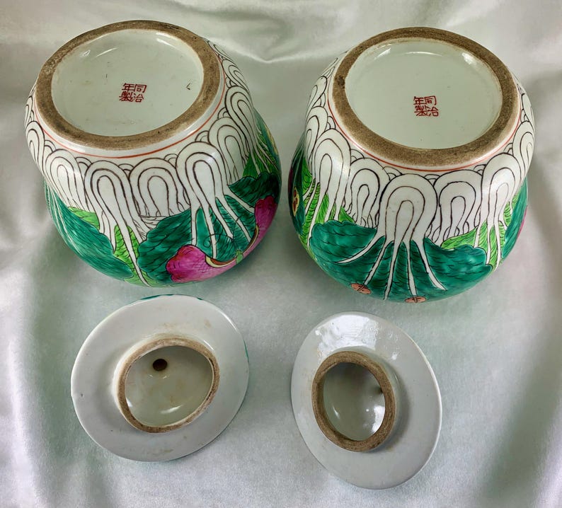 May include: Two antique, hand-painted ceramic jars with lids. The jars are decorated with green, white, and pink floral designs. The lids are white with a brown rim. The jars have a Chinese inscription on the bottom.