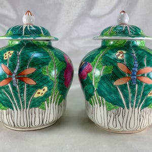 May include: Two matching ceramic jars with lids. The jars are painted with a green lotus leaf design, featuring orange dragonflies and pink and yellow flowers. The lids are green with white and red accents.