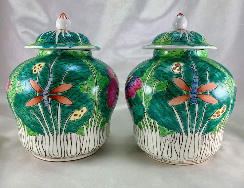 May include: Two matching ceramic jars with lids. The jars are painted with a green lotus leaf design, featuring orange dragonflies and pink and yellow flowers. The lids are green with white and red accents.
