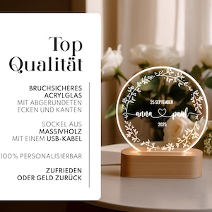May include: A personalized LED night light with a wooden base. The round, clear acrylic light features the names "anna & paul", the date "25 September 2025", and a floral wreath design. The text "Top Qualit&auml;t" is also visible.
