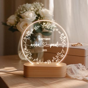 May include: A personalized wedding night light with a clear, circular design. The light features the names "anna & paul", the date "25 SEPTEMBER 2025", and a heart symbol, all within a floral wreath. It sits on a wooden base.