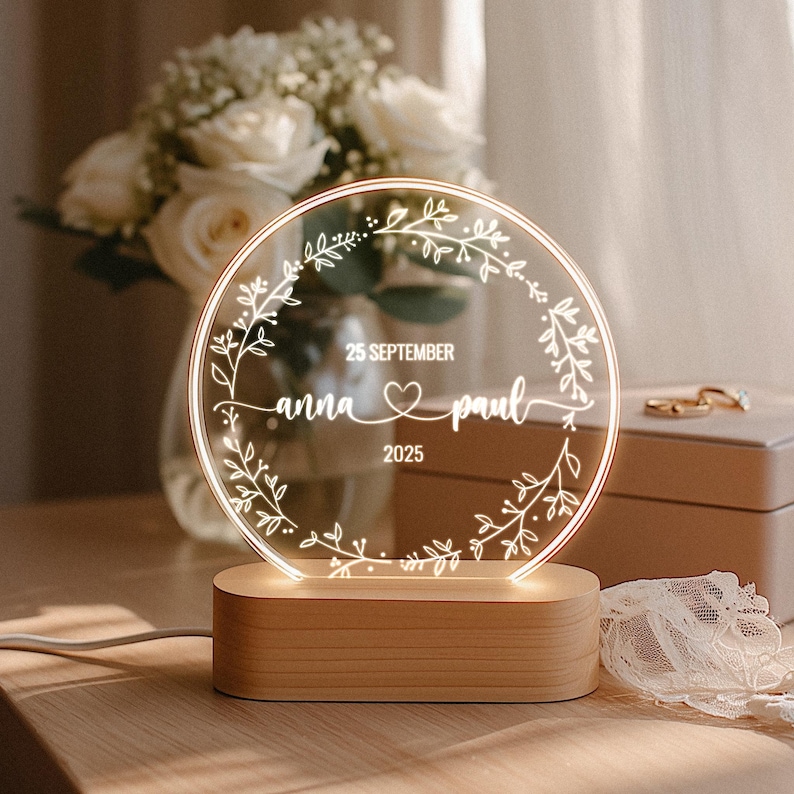 May include: A personalized wedding night light with a clear, circular design. The light features the names "anna & paul", the date "25 SEPTEMBER 2025", and a heart symbol, all within a floral wreath. It sits on a wooden base.