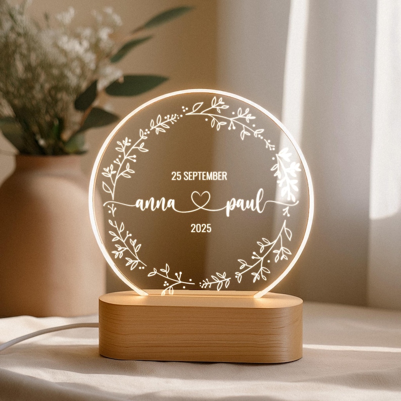 May include: A personalized wedding night light with a round, illuminated design. The clear acrylic panel features a floral wreath and the names "Anna & Paul" with the date "25 September 2025". The light sits on a rectangular wooden base.