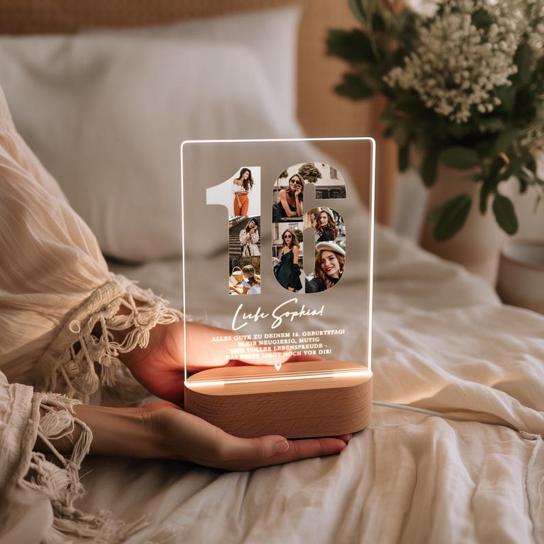 May include: A clear acrylic LED lamp with the number "16" cut out, filled with photos. The lamp sits on a wooden base and has the text "Liebe Sophia" and other text below. The lamp is held in someone's hands.