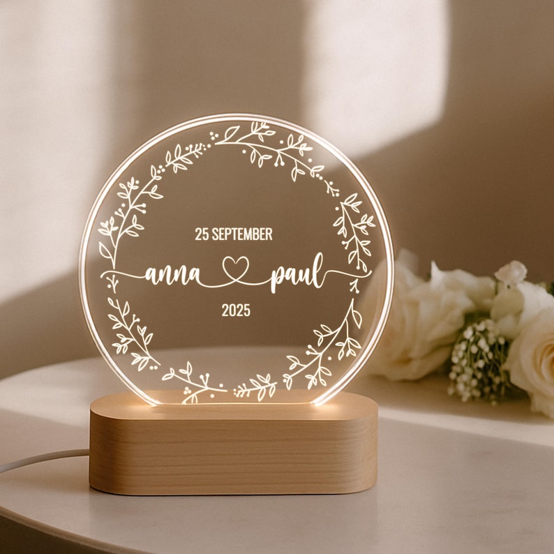 May include: A round, illuminated acrylic wedding sign with the names "anna & paul" and the date "25 September 2025" displayed. The sign is encircled by a floral wreath and sits on a wooden base.