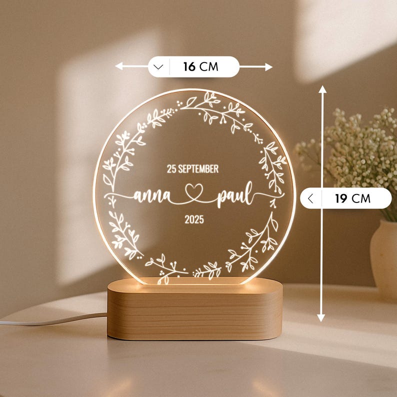 May include: A personalized LED night light, 19 cm tall and 16 cm wide, with a wooden base. The clear acrylic circle features a floral wreath design and the names "anna & paul" with the date "25 SEPTEMBER 2025".