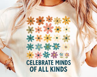 May include: Cream-colored t-shirt featuring a grid of colorful retro flowers. The shirt has the text "CELEBRATE MINDS OF ALL KINDS" in a teal font. The flowers are in shades of orange, pink, yellow, green, and blue.