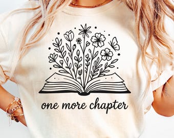 May include: Cream-coloured t-shirt featuring a black floral design emerging from an open book. The words "one more chapter" are printed below the book. The shirt is paired with blue jeans.