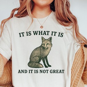 Op de afbeelding: Cr&egrave;mekleurig T-shirt met een gedetailleerde illustratie van een vos. De tekst "IT IS WHAT IT IS" staat boven de vos en "AND IT IS NOT GREAT" eronder, allemaal in donkergroene letters. Het shirt heeft korte mouwen.
