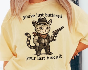 May include: Light yellow t-shirt featuring a cartoon cat dressed as a cowboy, holding a gun. The cat wears a hat, vest, and bandana. Text on the shirt reads "you've just buttered your last biscuit."