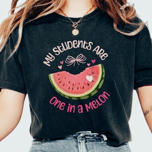 May include: Black t-shirt with the text "My Students Are One In A Melon" in pink lettering. A watermelon slice with a pink bow and hearts is featured on the shirt.