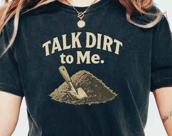 Peut inclure: T-shirt gris foncé avec l'inscription "TALK DIRT to Me." en police serif. En dessous, un dessin d'un tas de terre et d'une pelle. Le t-shirt est de style col rond.
