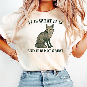 Op de afbeelding: Cr&egrave;mekleurig T-shirt met een groene illustratie van een vos en de tekst "IT IS WHAT IT IS" erboven en "AND IT IS NOT GREAT" eronder. De vos is afgebeeld in een gedetailleerde, vintage stijl.