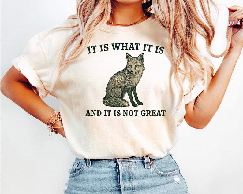 Op de afbeelding: Cr&egrave;mekleurig T-shirt met een groene illustratie van een vos en de tekst "IT IS WHAT IT IS" erboven en "AND IT IS NOT GREAT" eronder. De vos is afgebeeld in een gedetailleerde, vintage stijl.