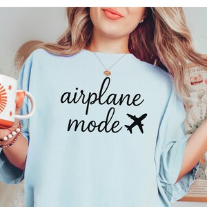 K&ouml;nnte beinhalten: Hellblaues T-Shirt mit den Worten "airplane mode" in schwarzer Schrift und einer schwarzen Flugzeuggrafik. Eine wei&szlig;e Tasse mit einem orangefarbenen Sonnendesign wird in der linken Hand gehalten.
