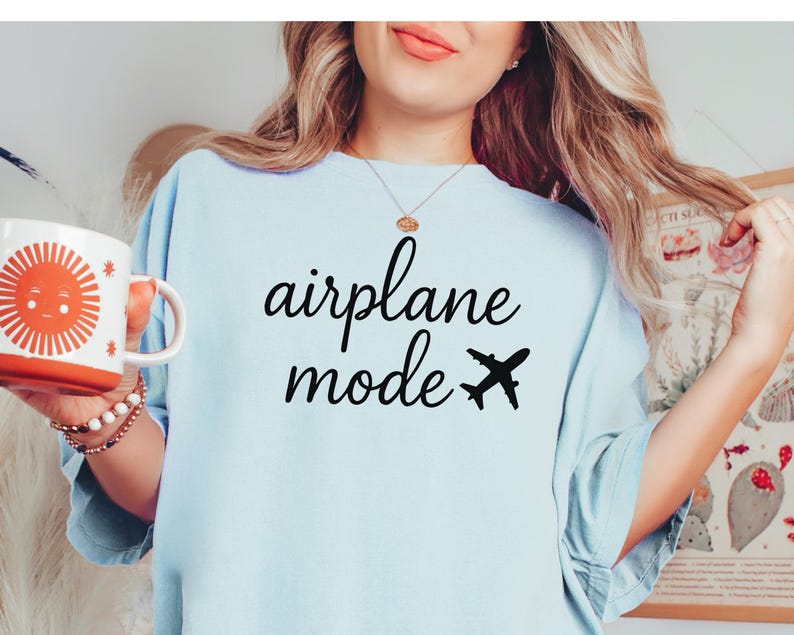 K&ouml;nnte beinhalten: Hellblaues T-Shirt mit den Worten "airplane mode" in schwarzer Schrift und einer schwarzen Flugzeuggrafik. Eine wei&szlig;e Tasse mit einem orangefarbenen Sonnendesign wird in der linken Hand gehalten.
