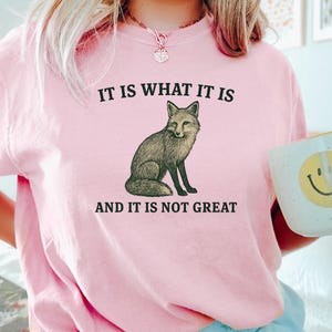 Op de afbeelding: Roze sweatshirt met de tekst "IT IS WHAT IT IS AND IT IS NOT GREAT" en een illustratie van een vos. Een witte mok met een gele smiley wordt in de hand gehouden.
