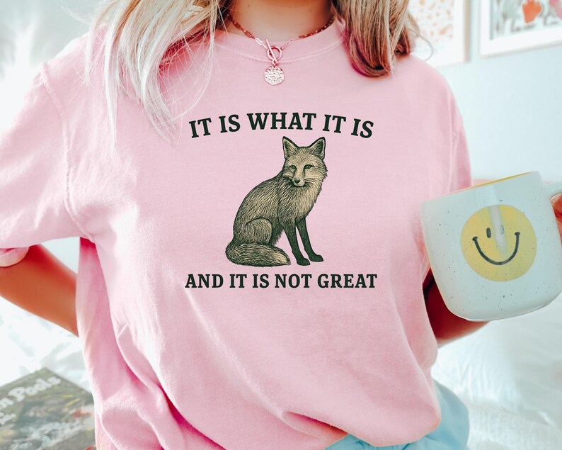 Op de afbeelding: Roze sweatshirt met de tekst "IT IS WHAT IT IS AND IT IS NOT GREAT" en een illustratie van een vos. Een witte mok met een gele smiley wordt in de hand gehouden.