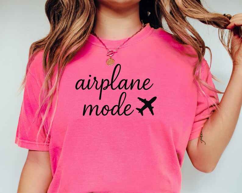 K&ouml;nnte beinhalten: Ein leuchtend pinkfarbenes T-Shirt mit dem schwarzen Schriftzug "airplane mode" und einer kleinen Flugzeug-Grafik. Das Shirt ist einfarbig und hat kurze &Auml;rmel.