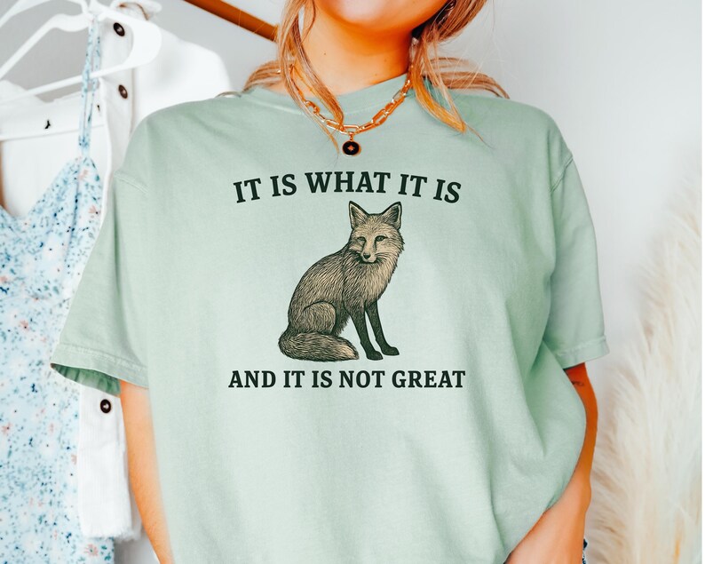 Op de afbeelding: Lichtgroen T-shirt met de tekst "IT IS WHAT IT IS AND IT IS NOT GREAT" en een gedetailleerde illustratie van een vos. Het shirt heeft korte mouwen en een ronde hals.