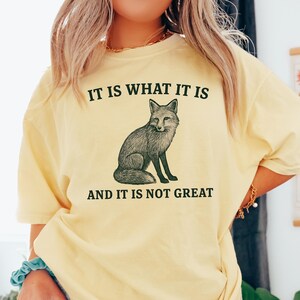 Op de afbeelding: Lichtgeel T-shirt met de tekst "IT IS WHAT IT IS" boven een gedetailleerde illustratie van een vos. Onder de vos staat de tekst "AND IT IS NOT GREAT". Het shirt heeft korte mouwen.