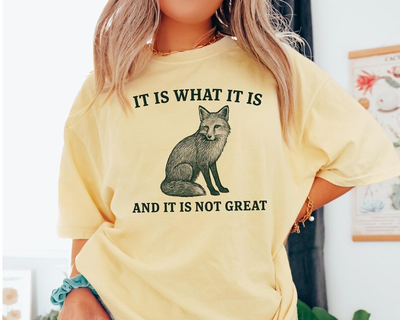 Op de afbeelding: Lichtgeel T-shirt met de tekst "IT IS WHAT IT IS" boven een gedetailleerde illustratie van een vos. Onder de vos staat de tekst "AND IT IS NOT GREAT". Het shirt heeft korte mouwen.